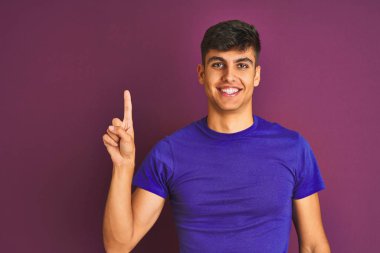 İzole edilmiş mor arka plan üzerinde duran tişört giyen genç hintli adam, kendinden emin ve mutlu bir şekilde gülümserken bir numaralı parmakla işaret ediyor..