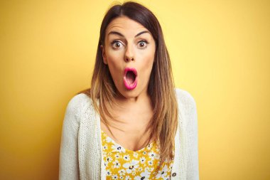 Sarı izole arka plan üzerinde duran ceket giyen genç güzel kadın korkmuş ve sürpriz ifade ile şok, korku ve heyecanlı yüz.
