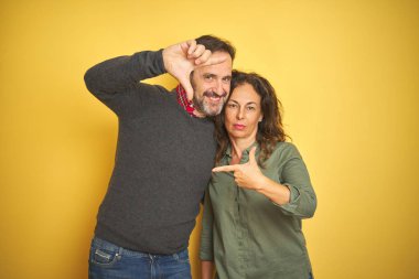 İzole sarı arka plan üzerinde güzel orta yaş çift mutlu yüz ile eller ve parmaklar ile çerçeve yapma gülümseyen. Yaratıcılık ve Fotoğrafçılık konsepti.