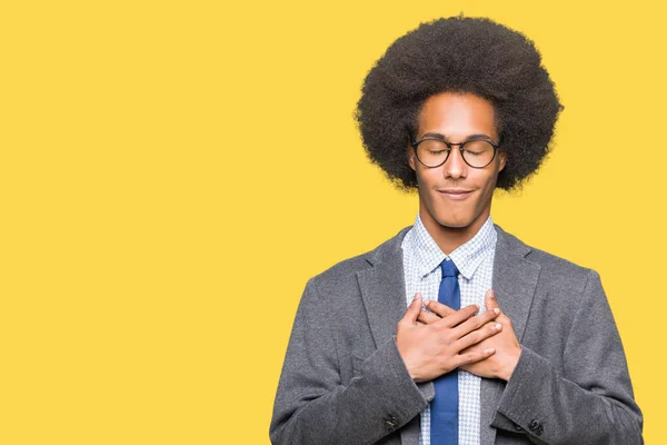 Young african american business man with afro hair wearing glasses smiling with hands on chest with closed eyes and grateful gesture on face. Health concept.