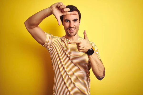 年轻的英俊男子与纹身穿着条纹T恤在孤立的黄色背景微笑 用手和手指与快乐的脸框架 创意与摄影理念 — 图库照片