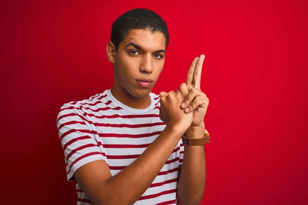 Young handsome arab man wearing striped t-shirt over isolated red background Holding symbolic gun with hand gesture, playing killing shooting weapons, angry face