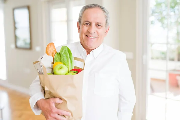 Handsome senior man holding paper bag full of fresh groceries and smiling at home