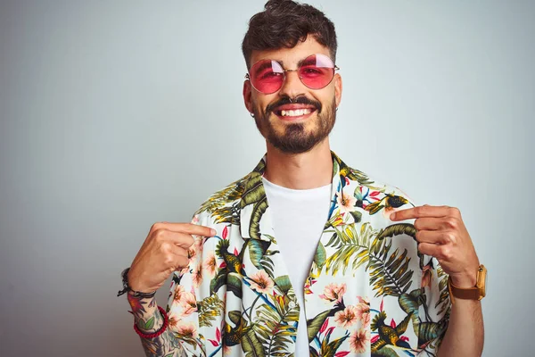 Man with tattoo on vacation wearing summer shirt sunglasses over isolated white background looking confident with smile on face, pointing oneself with fingers proud and happy.