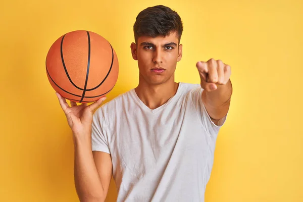年轻的印度运动员拿着篮球站在孤立的黄色背景上 用手指指着镜头 手签名 积极和自信的姿态从前面 — 图库照片