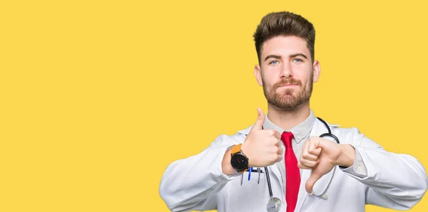 Young handsome doctor man wearing medical coat Doing thumbs up and down, disagreement and agreement expression. Crazy conflict