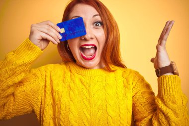 Sarı izole arka plan üzerinden kredi kartı tutan genç güzel Redhead kadın çok mutlu ve heyecanlı, kazanan ifade zafer büyük gülümseme ile çığlık kutluyor ve eller kaldırdı