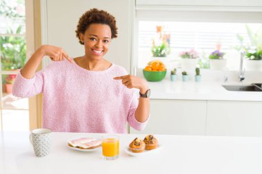 Genç Afrikalı Amerikan kadın evde sabah kahvaltı yeme yüzünde gülümseme ile kendine güvenen görünümlü, parmaklarıyla gurur ve mutlu kendini işaret.