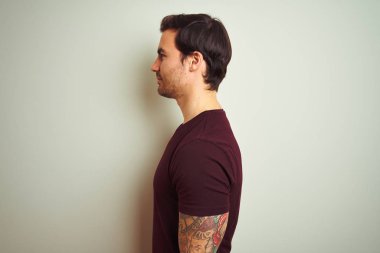 Yüze bakarak izole beyaz arka plan üzerinde mor rahat t-shirt giyen dövme ile genç yakışıklı adam, güvenli bir gülümseme ile doğal yüzü ile profil poz rahatlayın.