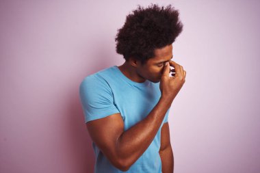 Afro saç ile Afrika Amerikan adam izole pembe arka plan üzerinde duran mavi t-shirt ayakta sürtünme burun ve gözleri duygu yorgunluk ve baş ağrısı yorgun. Stres ve hayal kırıklığı kavramı.