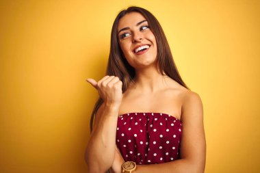 İzole sarı arka plan üzerinde duran kırmızı t-shirt giyen genç güzel kadın mutlu yüz görünümlü ve başparmak ile yan işaret gülümseyerek.