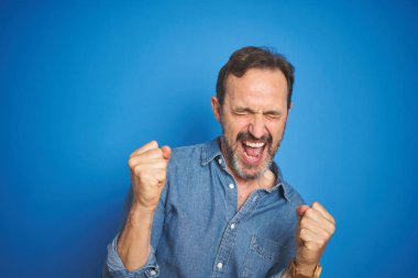 İzole mavi arka plan üzerinde gri saçlı yakışıklı Orta Çağ kıdemli adam çok mutlu ve heyecanlı silah ile kazanan jest yaparak, gülümseyen ve başarı için çığlık kaldırdı. Kutlama konsepti.