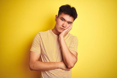 İzole sarı arka plan üzerinde ayakta tişört giyen genç Asyalı Çinli adam yorgun bakıyor ve çapraz kolları ile depresyon sorunları ile sıkılmış.