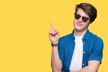 Kameraya bakarak yan el ve parmak ile işaret eden genç yakışıklı adam yüzünde kocaman bir gülümseme ile izole arka plan üzerinde güneş gözlüğü takıyor.