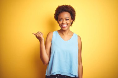 Güzel Afrikalı Amerikan kadın izole sarı arka plan üzerinde mutlu yüz görünümlü ve başparmak yukarı ile yan işaret gülümseyen üzerinde zarif gömlek giyiyor.