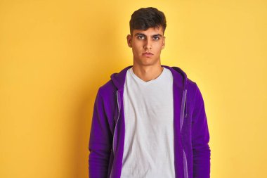 İzole sarı arka plan üzerinde duran mor sweatshirt giyen genç hintli adam yüzünde ciddi ifade ile rahat. Kameraya basit ve doğal bir bakış.