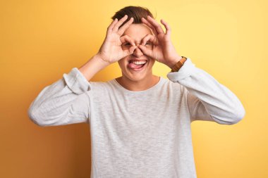 Beyaz rahat t-shirt giyen genç yakışıklı adam izole sarı arka plan üzerinde duran dürbün dil dışarı yapışmasını gibi ok jest yapıyor, gözleri parmaklardan bakarak. Çılgın ifade.