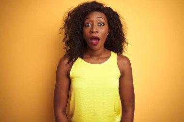 İzole sarı arka plan üzerinde duran t-shirt giyen genç Afrikalı-Amerikalı kadın korkmuş ve sürpriz ifade ile şok, korku ve heyecanlı yüz.