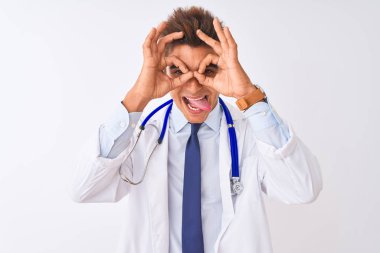 Genç yakışıklı doktor adam izole beyaz arka plan üzerinde stetoskop giyen dürbün dil dışarı yapışmasını gibi ok jest yapıyor, gözleri parmaklardan bakarak. Çılgın ifade.