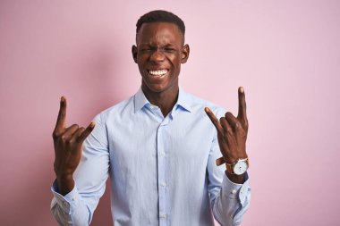 Mavi zarif gömlek giyen Afrika Amerikan adam izole pembe arka plan üzerinde ayakta çılgın ifade ile eller yukarı rock sembolü yapıyor bağırıyor. Müzik yıldızı. Ağır kavram.