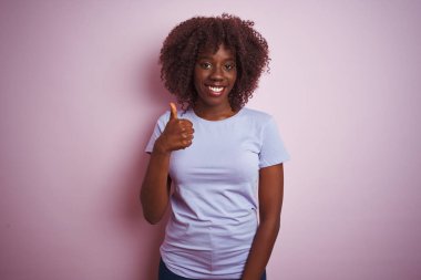 Genç Afrikalı Afro kadın el ile jest kadar mutlu başparmak yapıyor izole pembe arka planda ayakta tişört giyiyor. Başarı göstererek kameraya bakarak ifadeyi onaylama.