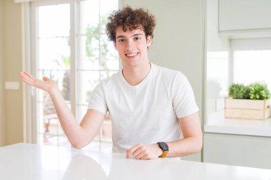 Evde beyaz t-shirt giyen genç yakışıklı adam neşeli sunumu gülümseyen ve kamera bakarak el avuç ile işaret.