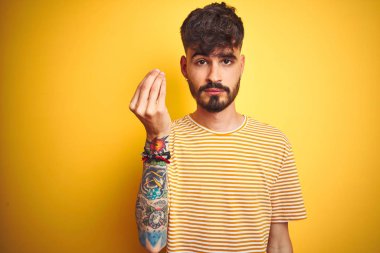 Dövme ile genç adam izole sarı arka plan üzerinde ayakta çizgili t-shirt dikili el ve parmaklar ile Italyan jest yapıyor emin ifade
