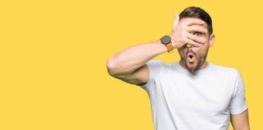 Yakışıklı adam utanç ifadesi ile parmaklarımın arasından seyir yüz ve gözler el ile kapsayan şok bakmak rahat beyaz t-shirt giyen.