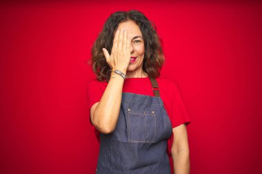 Orta yaş kıdemli kadın el ile bir göz kapsayan kırmızı izole arka plan üzerinde apron üniforma giyiyor, yüz ve sürpriz duygu kendine güvenen gülümseme.