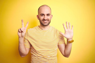 Sarı izole arka plan üzerinde rahat çizgili t-shirt giyen ve kendinden emin ve mutlu gülümseyen iken parmaklarının sayısı yedi ile gösteren sakal ile genç kel adam.
