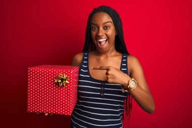 Genç afro-amerikan kadın izole kırmızı arka plan üzerinde duran doğum günü hediyesi tutan çok mutlu el ve parmak ile işaret