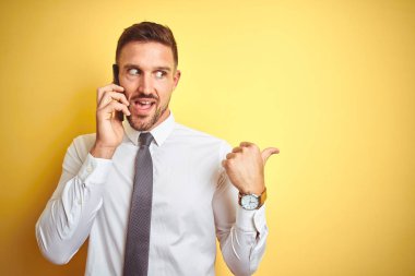 Sarı izole arka plan üzerinde telefonda konuşurken genç yakışıklı iş adam işaret ve mutlu yüz gülümseyen yan başparmak ile gösteren