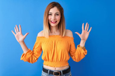 Turuncu rahat t-shirt giyen güzel redhead kadın izole mavi arka plan üzerinde duran gösteren ve emin ve mutlu gülümserken parmakları on numara ile işaret.