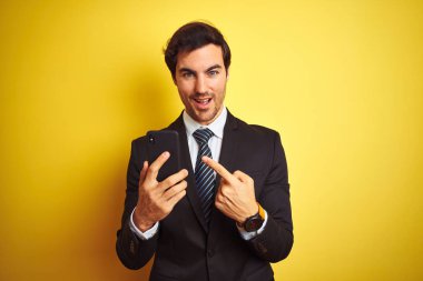 El ve parmak ile işaret çok mutlu izole sarı arka planda ayakta akıllı telefon kullanarak genç yakışıklı işadamı