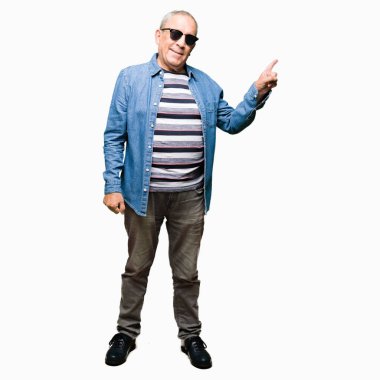 Denim ceket ve güneş gözlüğü giyen yakışıklı yaşlı adam yüzünde büyük bir gülümseme ile, kameraya bakarak yan el ve parmak ile işaret.