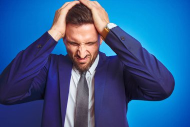 Mavi izole arka plan üzerinde genç yakışıklı iş adamın resim yakın baş ağrısı umutsuz ve vurguladı çünkü ağrı ve migren. Eller baş.