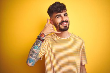 Dövme ile genç adam izole sarı arka plan üzerinde duran çizgili t-shirt ayakta telefonda konuşmak gibi el ve parmaklar ile telefon jest yapıyor gülümsüyor. İletişim kavramları.