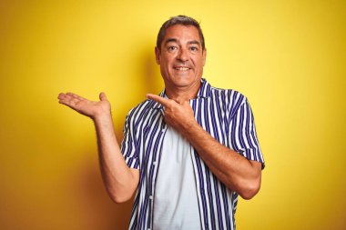 El ile sunarak ve parmak ile işaret ederken kamera gülümseyen izole sarı arka plan üzerinde ayakta çizgili gömlek giyen yakışıklı orta yaş adam.