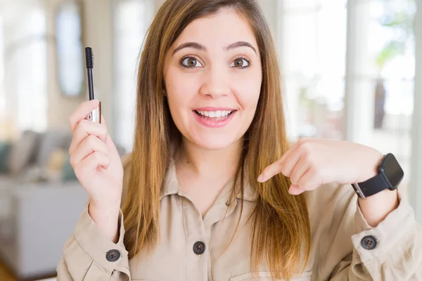 Beautiful young woman putting lash mascara on eyelashes with surprise face pointing finger to himself