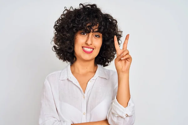 Young arab woman with curly hair wearing casual shirt over isolated white background smiling with happy face winking at the camera doing victory sign. Number two.