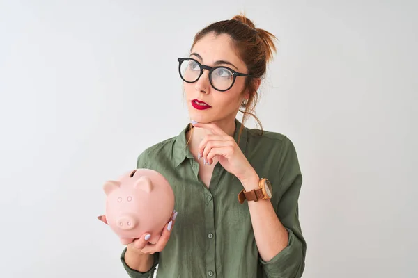 美丽的红发女人戴着眼镜抱着小猪银行在孤立的白色背景严肃面对思考问题 很困惑的想法 — 图库照片