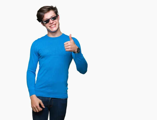 Young man wearing funny thug life glasses over isolated background doing happy thumbs up gesture with hand. Approving expression looking at the camera with showing success.