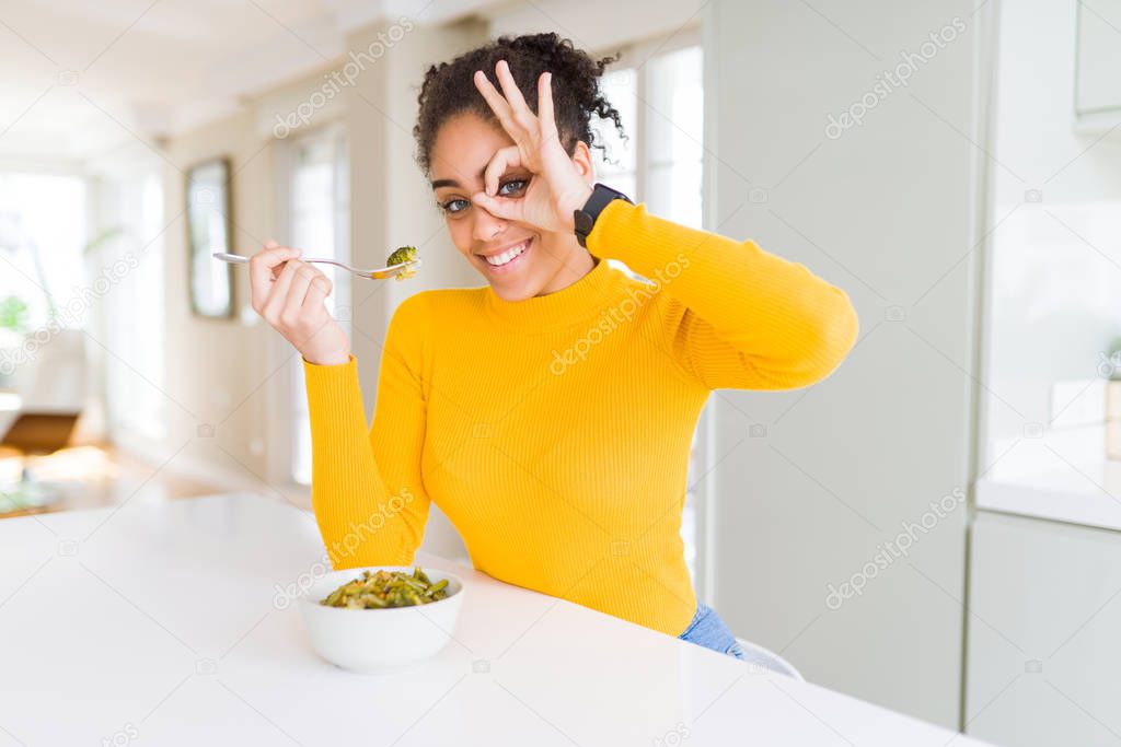 Young african american woman eating healthy green vegatables with happy face smiling doing ok sign with hand on eye looking through fingers