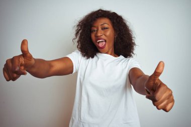 Genç afro-amerikan kadın izole beyaz arka plan üzerinde duran el ile olumlu jest yaptığını onaylayan t-shirt giyen, başparmak kadar gülümseyerek ve başarı için mutlu. Kazanan jest.