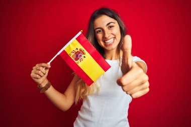 Kırmızı izole arka plan üzerinde İspanyol bayrağı tutan genç güzel kadın büyük bir gülümseme ile ok işareti, parmak yukarı başparmak, mükemmel işareti yapıyor mutlu