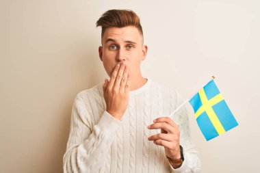 Genç yakışıklı adam izole beyaz arka plan kapağı ağız üzerinde sweadish İsveç bayrağı tutan el hata için utanç ile şok, korku ifadesi, sessizlik, gizli kavram korkmuş