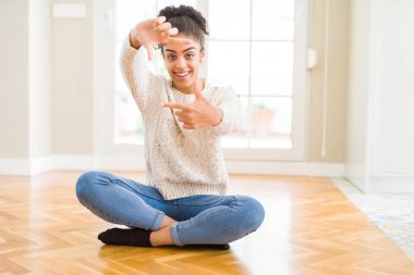 Afro saçlı güzel genç Afrikalı Amerikalı kadın, mutlu yüz ile eller ve parmaklar ile çerçeve yapma gülümseyen yerde oturuyor. Yaratıcılık ve Fotoğrafçılık konsepti.
