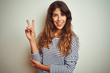 Beyaz izole arka plan üzerinde çizgili t-shirt giyen genç güzel kadın mutlu yüz zafer işareti yapıyor kamera göz kırpıyor ile gülümsüyor. İki numara.