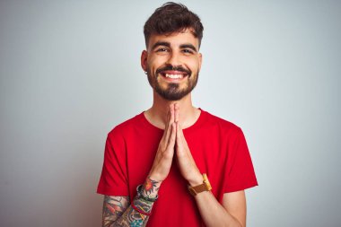 Dövme ile genç adam izole beyaz arka plan üzerinde duran kırmızı t-shirt ayakta affetmek için bağışlamak isteyen eller ile birlikte dua.