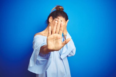 Genç güzel kadın mavi izole arka plan üzerinde topuz saç modeli giyen elleri ile gözleri kaplayan ve üzgün ve korku ifadesi ile stop jest yapıyor. Utanmış ve olumsuz kavram.